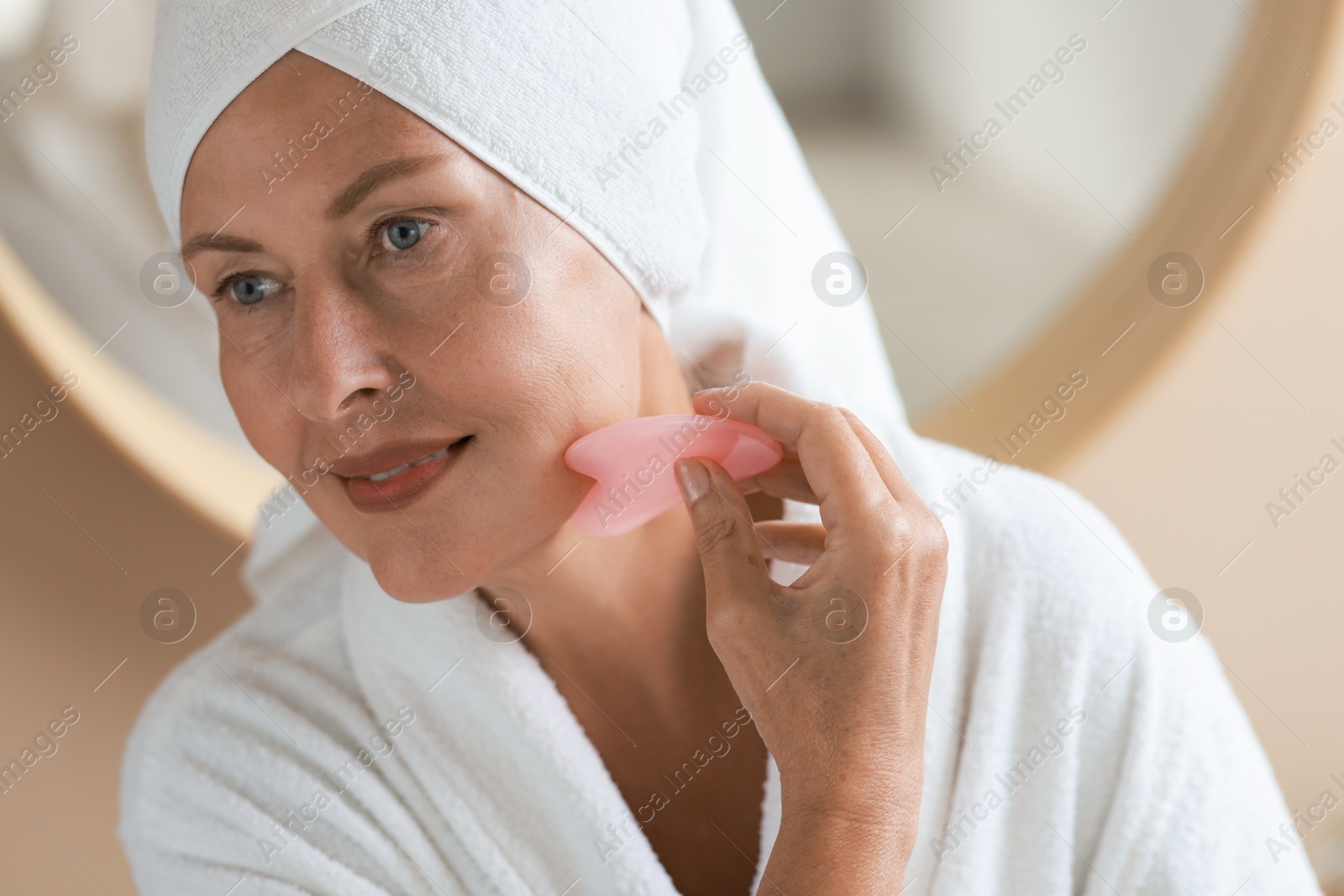 Photo of Beautiful young woman doing facial massage with gua sha tool indoors