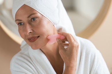 Beautiful young woman doing facial massage with gua sha tool indoors