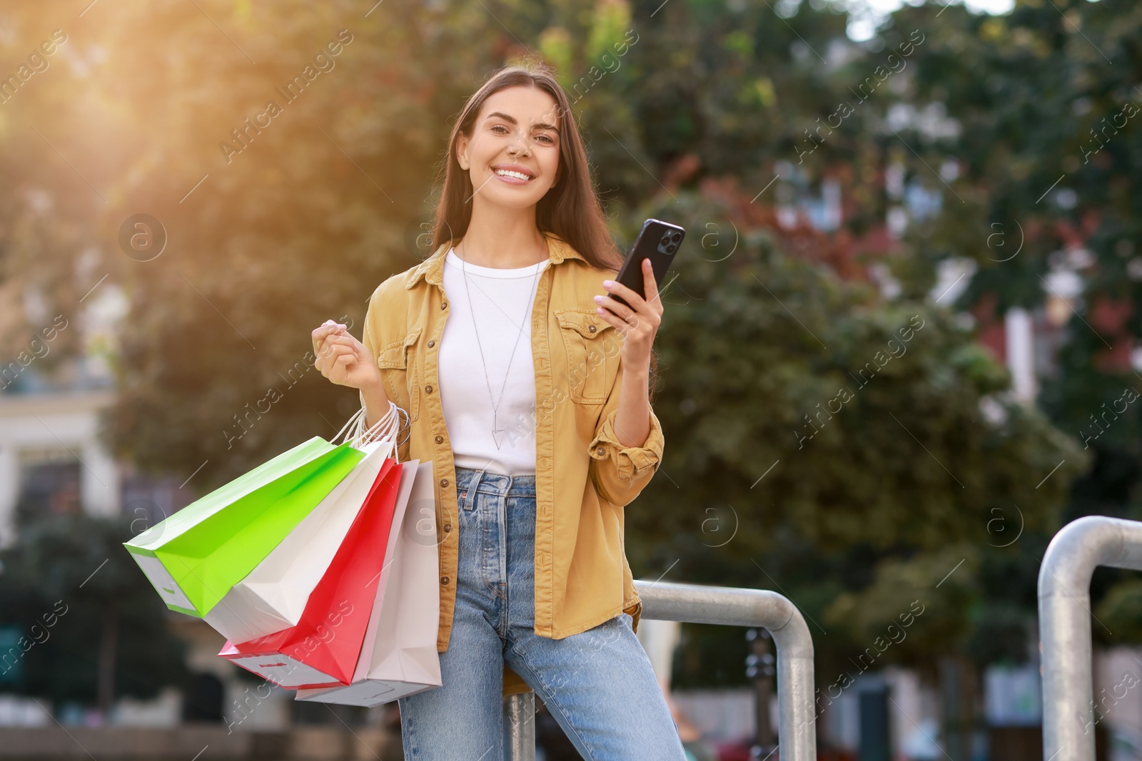 Photo of Happy woman with colorful shopping bags using smartphone outdoors