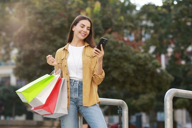 Photo of Happy woman with colorful shopping bags using smartphone outdoors, space for text