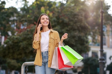 Photo of Happy woman with colorful shopping bags talking on smartphone outdoors