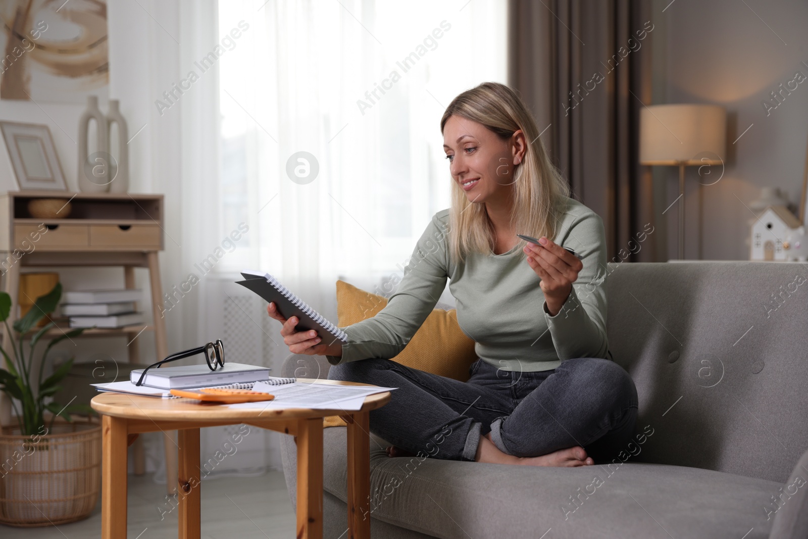 Photo of Budget planning. Woman with notebook and pen on sofa indoors