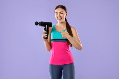 Young woman pointing at percussive massager on light purple background