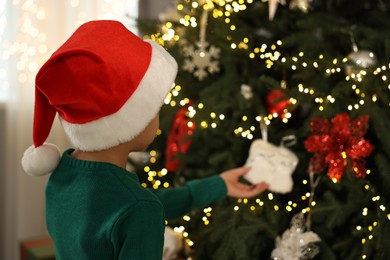 Cute little boy in Santa hat decorating Christmas tree at home