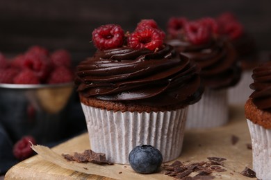 Photo of Tasty cupcakes with chocolate cream and raspberries on wooden board, closeup