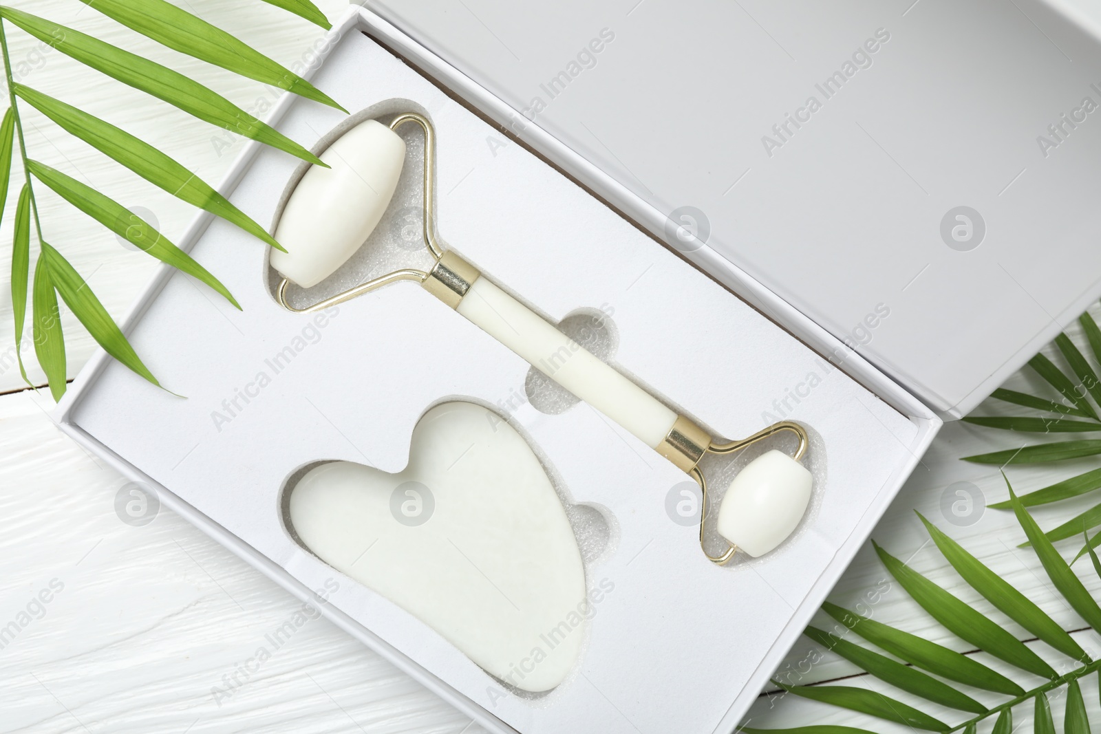 Photo of Face roller, gua sha tool in box and palm leaves on white wooden background, flat lay