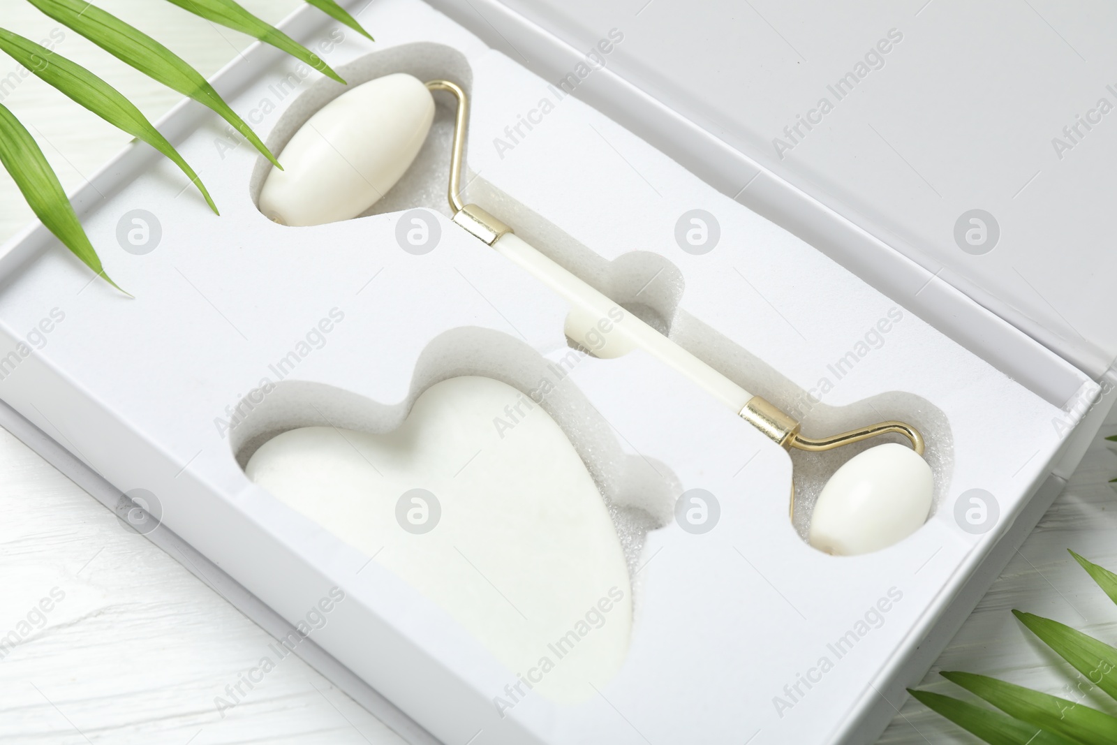 Photo of Face roller, gua sha tool in box and palm leaves on white wooden background, closeup