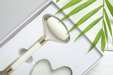 Photo of Face roller, gua sha tool in box and palm leaf on white wooden background, top view