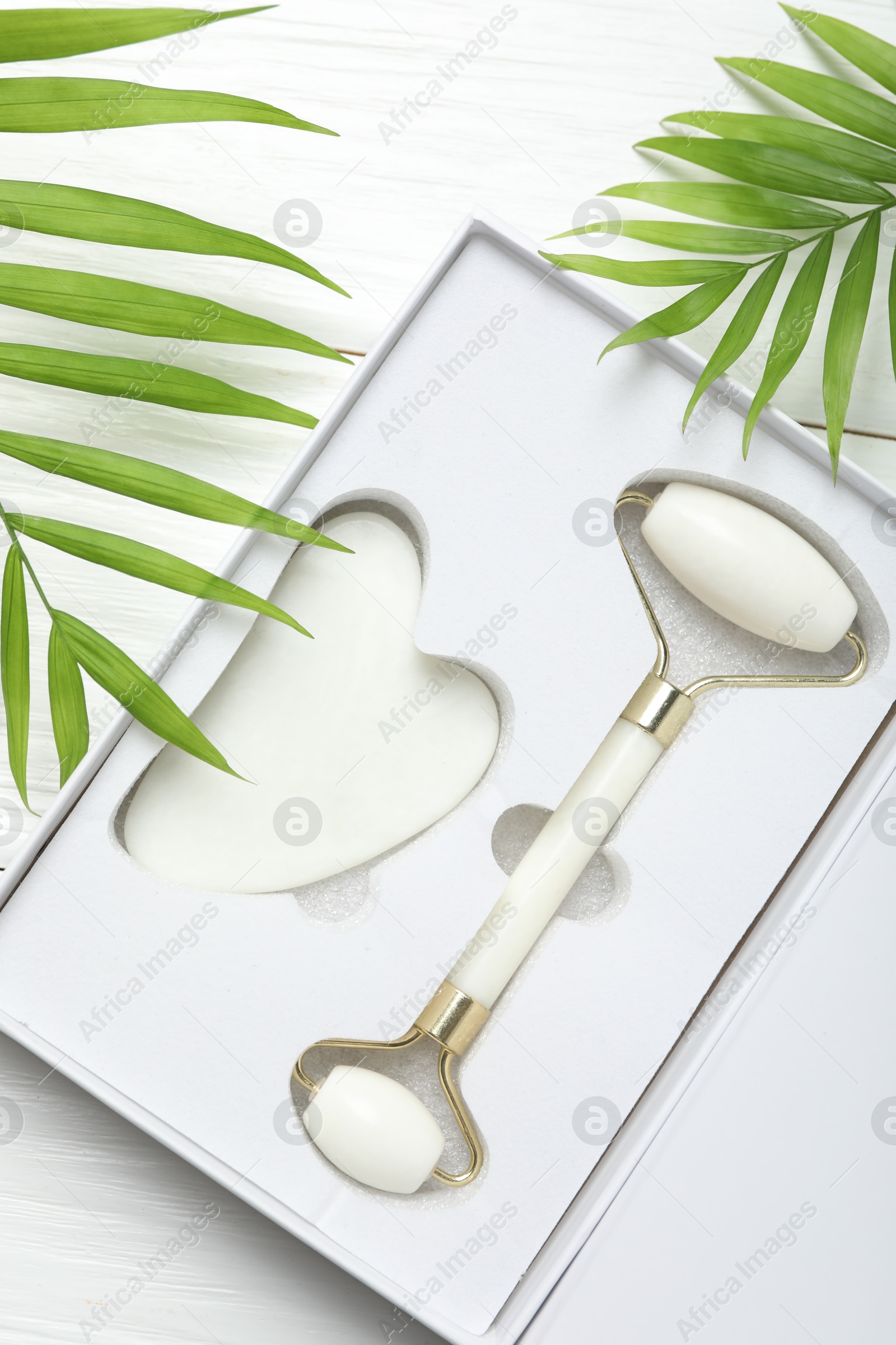 Photo of Face roller, gua sha tool in box and palm leaves on white wooden background, flat lay