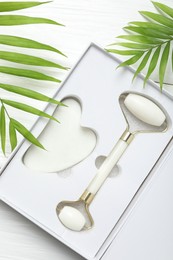 Photo of Face roller, gua sha tool in box and palm leaves on white wooden background, flat lay