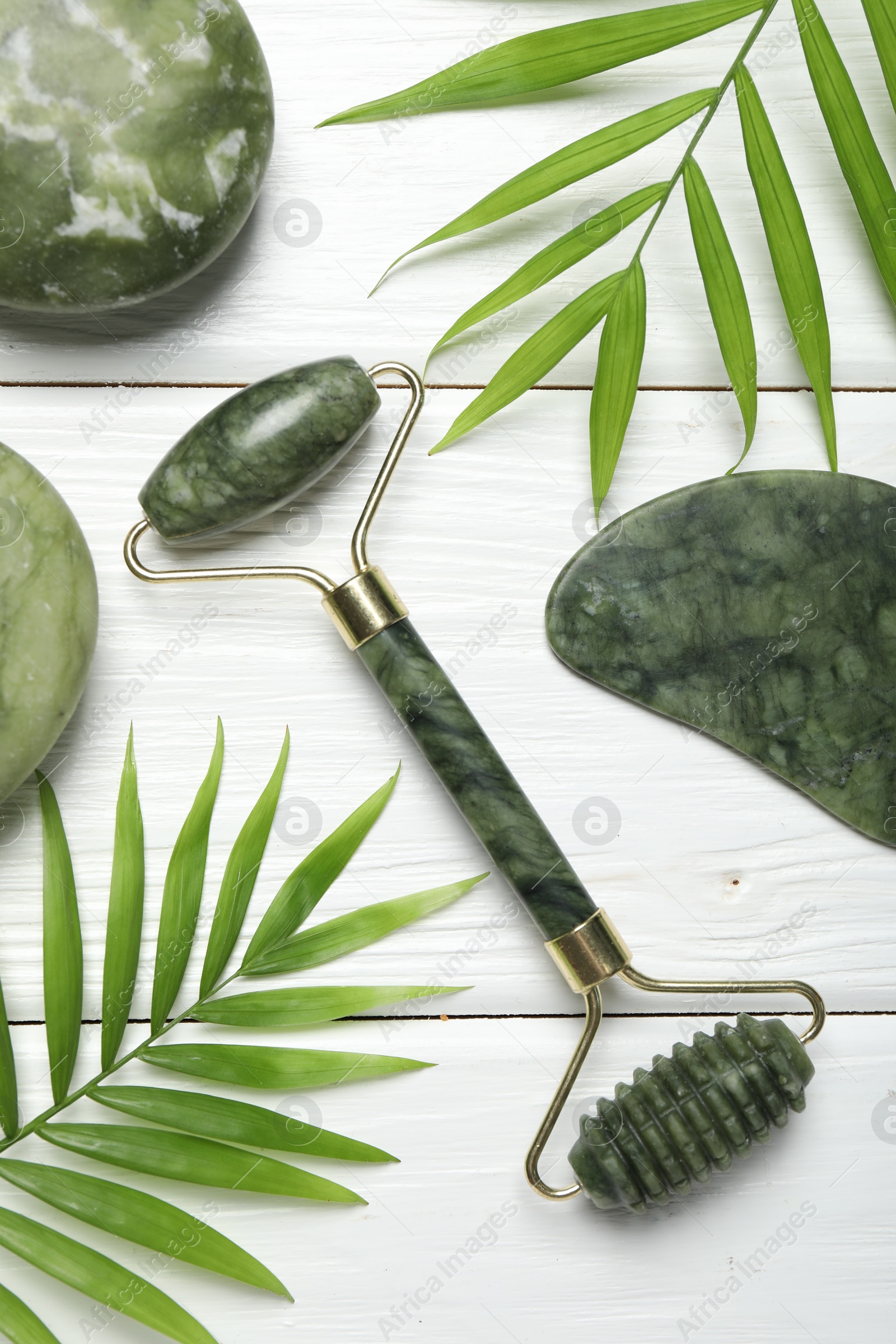 Photo of Face roller, gua sha tool, massage stones and palm leaves on white wooden background, flat lay