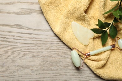 Photo of Face roller, gua sha tool, towel and ruscus branch on wooden background, flat lay. Space for text