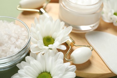 Photo of Face roller, gua sha tool, cosmetic product, sea salt and white chrysanthemum flowers on green background, closeup