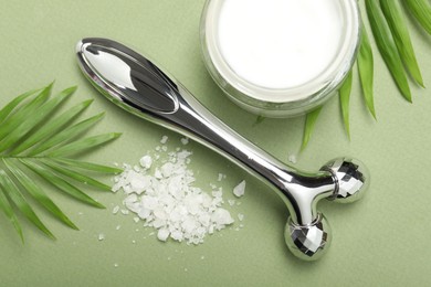 Photo of Metal face roller, cosmetic product, sea salt and palm leaves on green background, flat lay