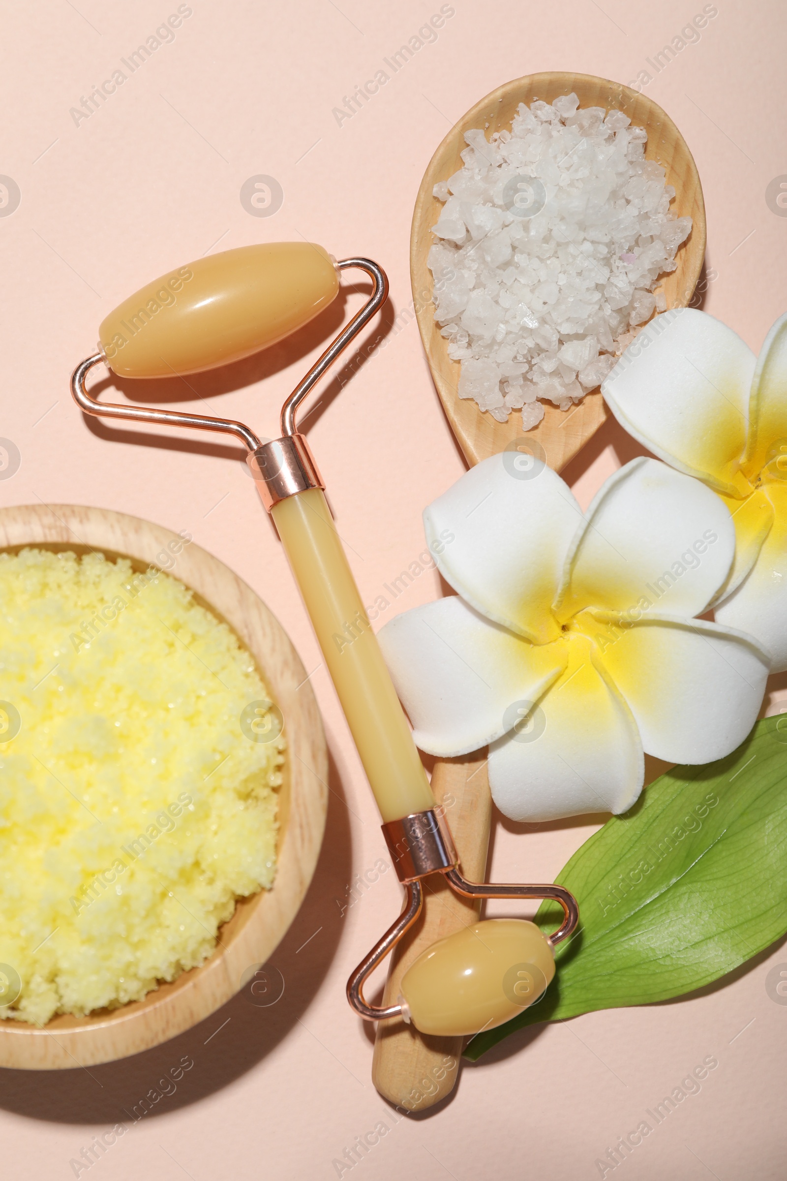 Photo of Face roller, body scrub, sea salt, plumeria flowers and green leaf on beige background, flat lay