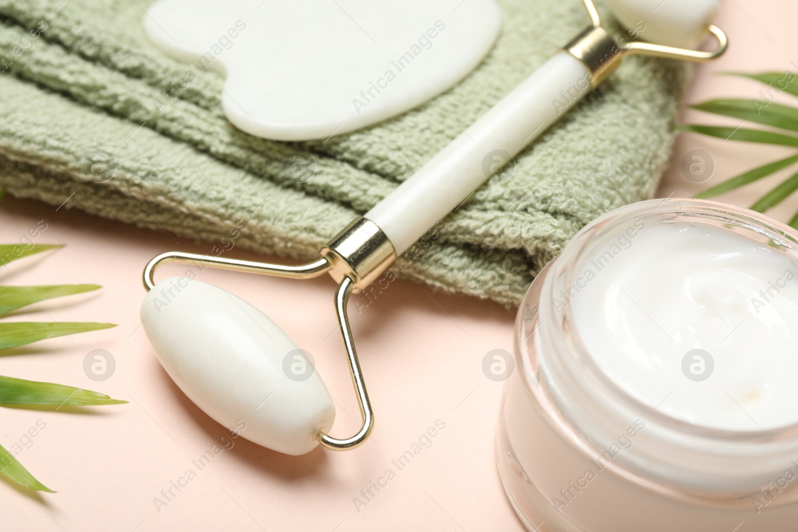 Photo of Face roller, gua sha tool, towel and cosmetic product on beige background, closeup