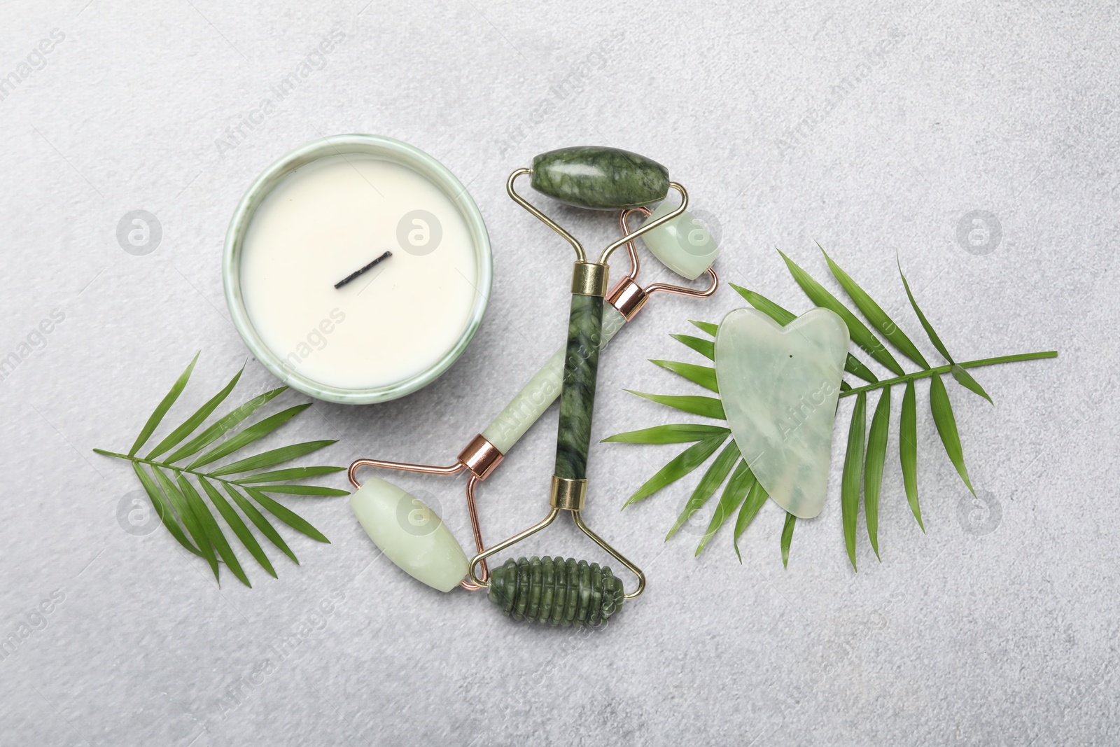 Photo of Face rollers, gua sha tool, scented candle and palm leaves on gray background, flat lay