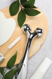 Metal face roller, gua sha tool, cosmetic products and green leaves on gray background, flat lay