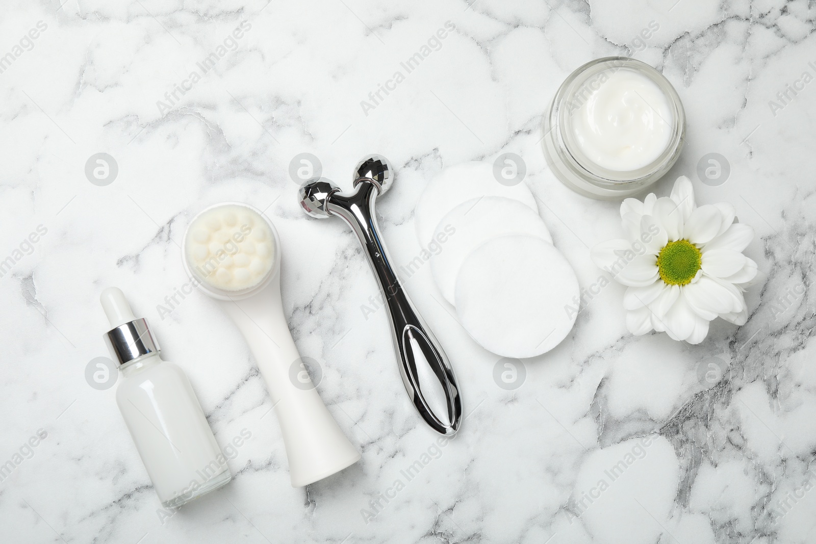 Photo of Metal face roller, cleansing brush, cosmetic products, cotton pads and chrysanthemum flower on white marble background, flat lay