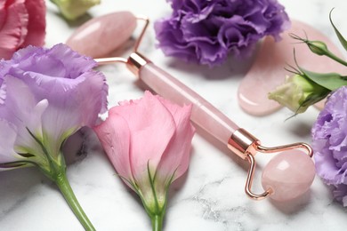 Photo of Face roller, gua sha tool and beautiful flowers on white marble background, closeup