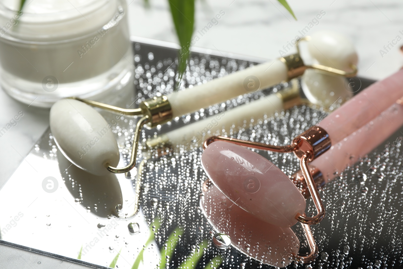 Photo of Face roller with water drops, cosmetic product and mirror on table, closeup