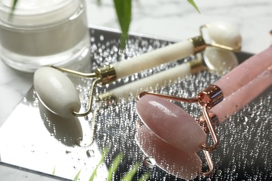 Photo of Face roller with water drops, cosmetic product and mirror on table, closeup