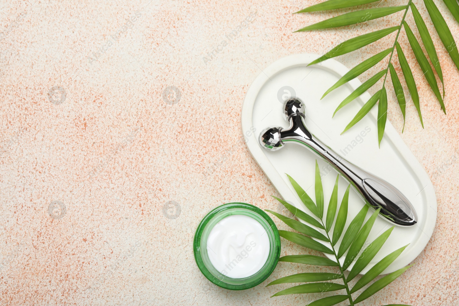 Photo of Metal face roller, cosmetic product and palm leaves on beige textured background, flat lay. Space for text