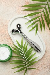 Photo of Metal face roller, cosmetic product and palm leaves on beige textured background, flat lay