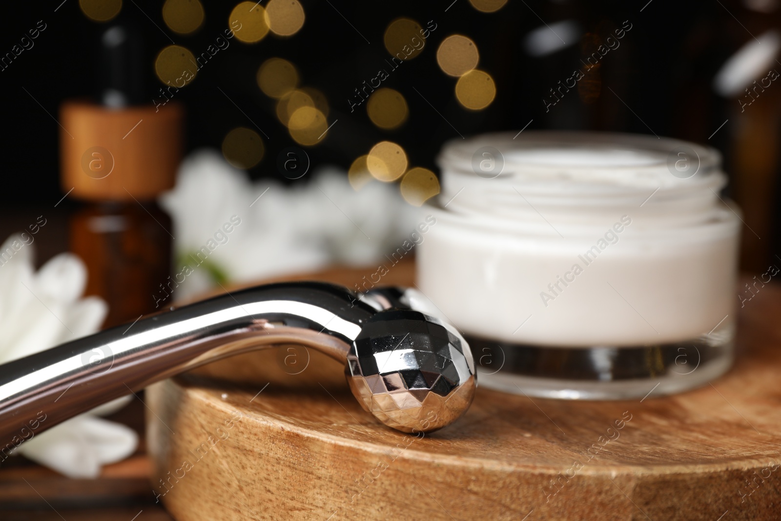 Photo of Metal face roller and cosmetic product on table against blurred lights, closeup