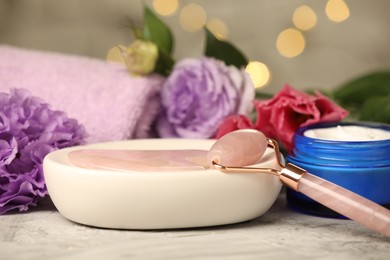 Face roller, gua sha tool, cosmetic product and beautiful flowers on gray textured table against blurred lights, closeup