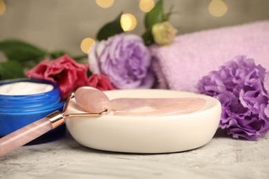 Photo of Face roller, gua sha tool, cosmetic product and beautiful flowers on gray textured table against blurred lights, closeup