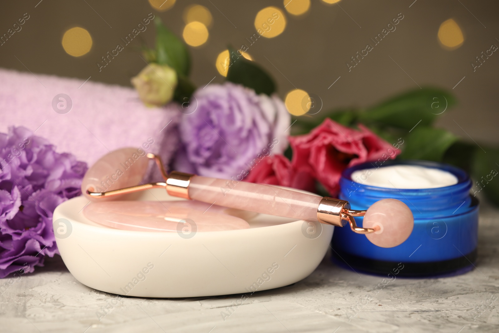 Photo of Face roller, gua sha tool, cosmetic product and beautiful flowers on gray textured table against blurred lights, closeup