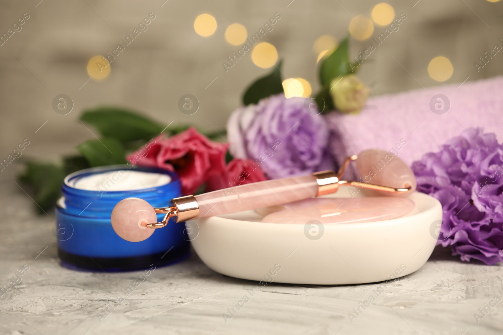 Photo of Face roller, gua sha tool, cosmetic product and beautiful flowers on gray textured table against blurred lights, closeup