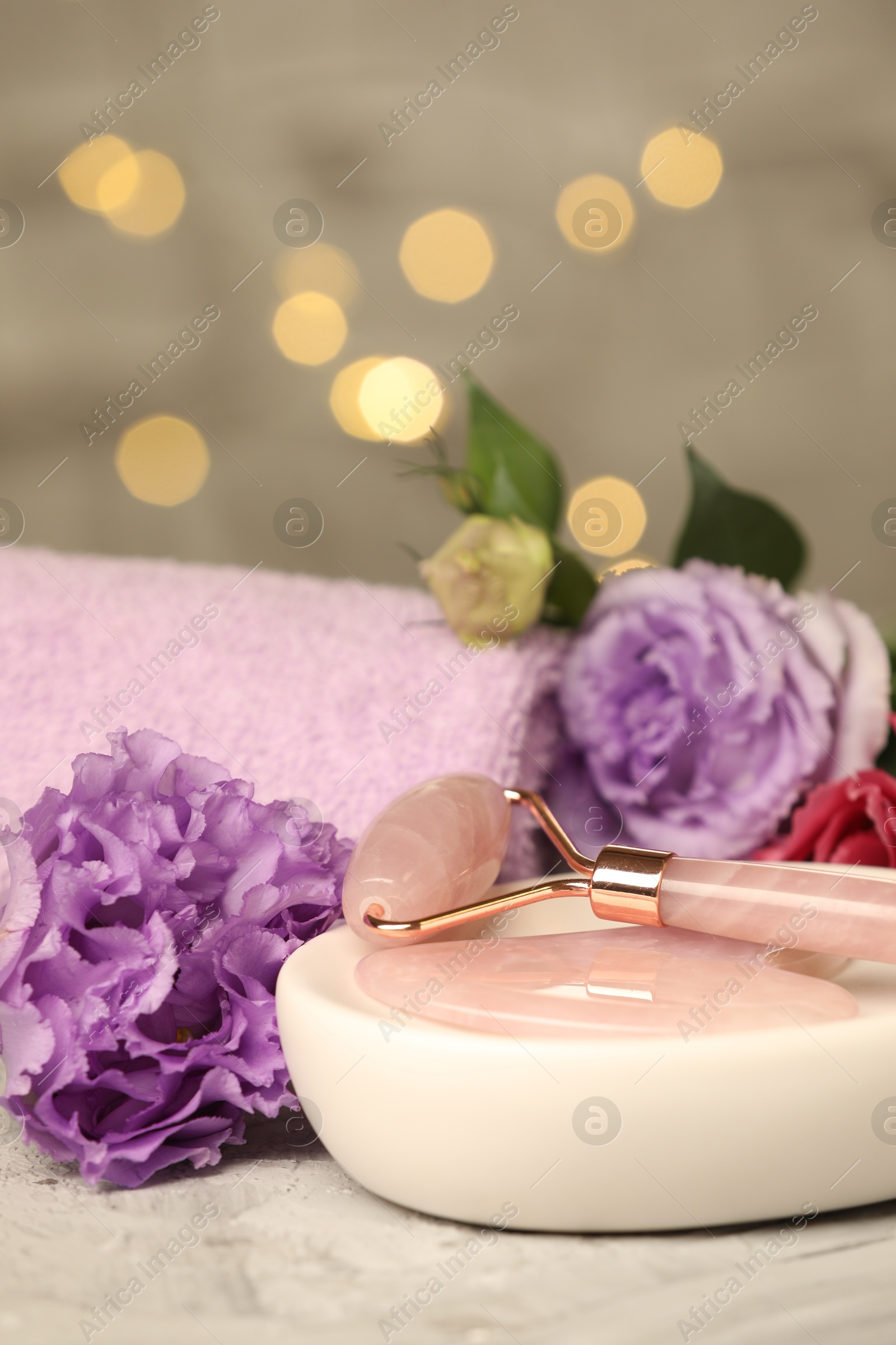 Photo of Face roller, gua sha tool, towel and beautiful flowers on gray textured table against blurred lights, closeup