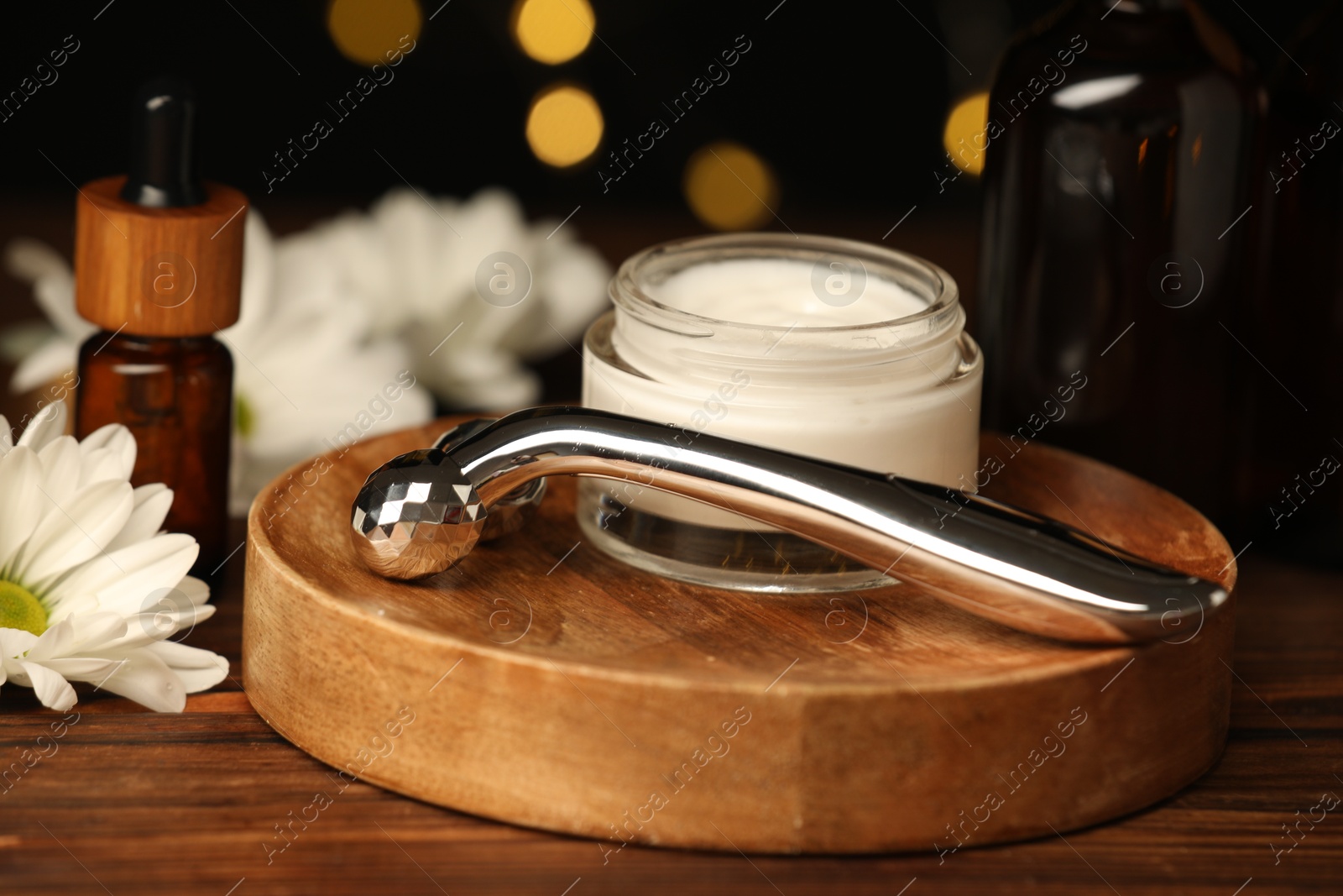 Photo of Metal face roller, cosmetic products and chrysanthemum flower on wooden table against blurred lights, closeup