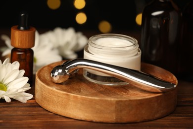 Metal face roller, cosmetic products and chrysanthemum flower on wooden table against blurred lights, closeup