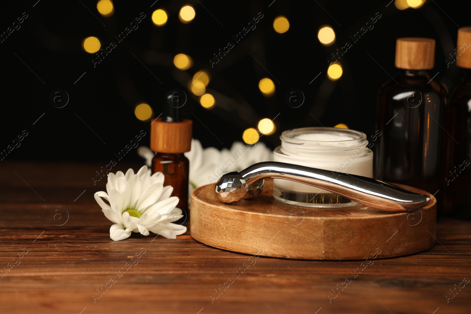 Photo of Metal face roller, cosmetic products and chrysanthemum flower on wooden table against blurred lights