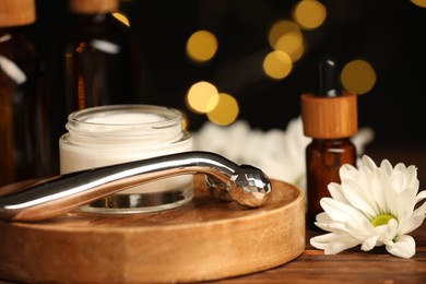 Metal face roller, cosmetic products and chrysanthemum flower on wooden table against blurred lights, closeup