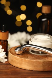 Metal face roller, cosmetic products and chrysanthemum flower on wooden table against blurred lights, closeup