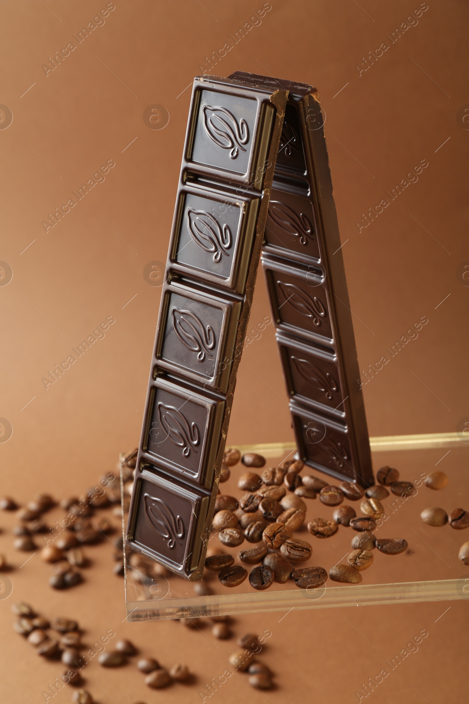 Photo of Pieces of tasty chocolate bar and coffee beans on glass surface against brown background