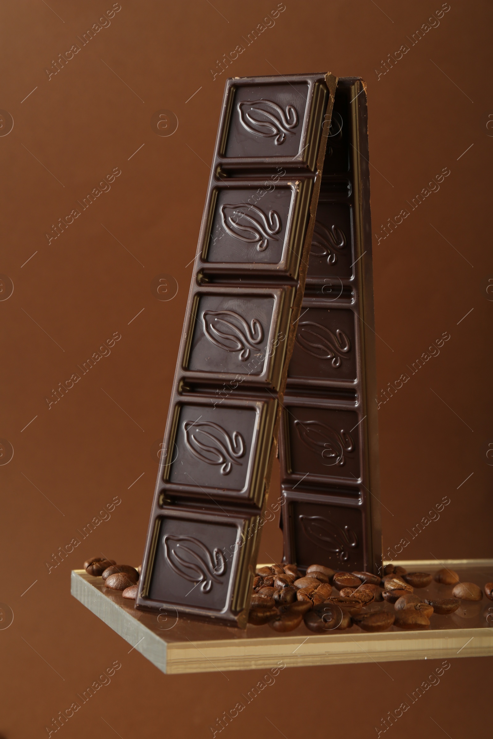 Photo of Pieces of tasty chocolate bar and coffee beans on glass surface against brown background