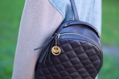 Photo of Woman with wooden keychain in shape of smiley face and black leather decor on her backpack outdoors, closeup