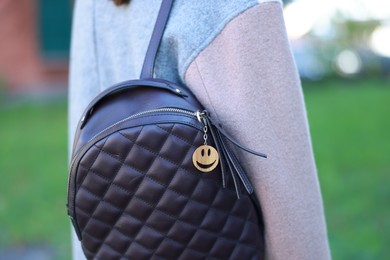 Photo of Woman with wooden keychain in shape of smiley face and black leather decor on her backpack outdoors, closeup