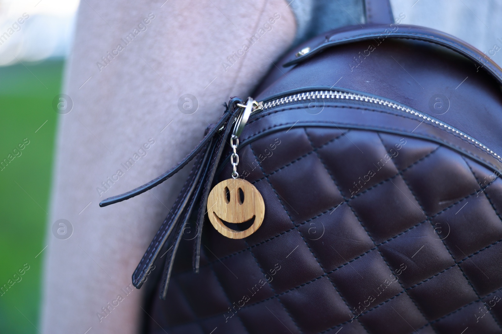 Photo of Woman with wooden keychain in shape of smiley face and black leather decor on her backpack outdoors, closeup. Selective focus