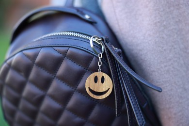 Photo of Woman with wooden keychain in shape of smiley face and black leather decor on her backpack outdoors, closeup. Selective focus