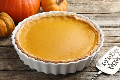 Photo of Tasty homemade pumpkin pie in baking dish, fresh pumpkins and cake server on wooden table, closeup