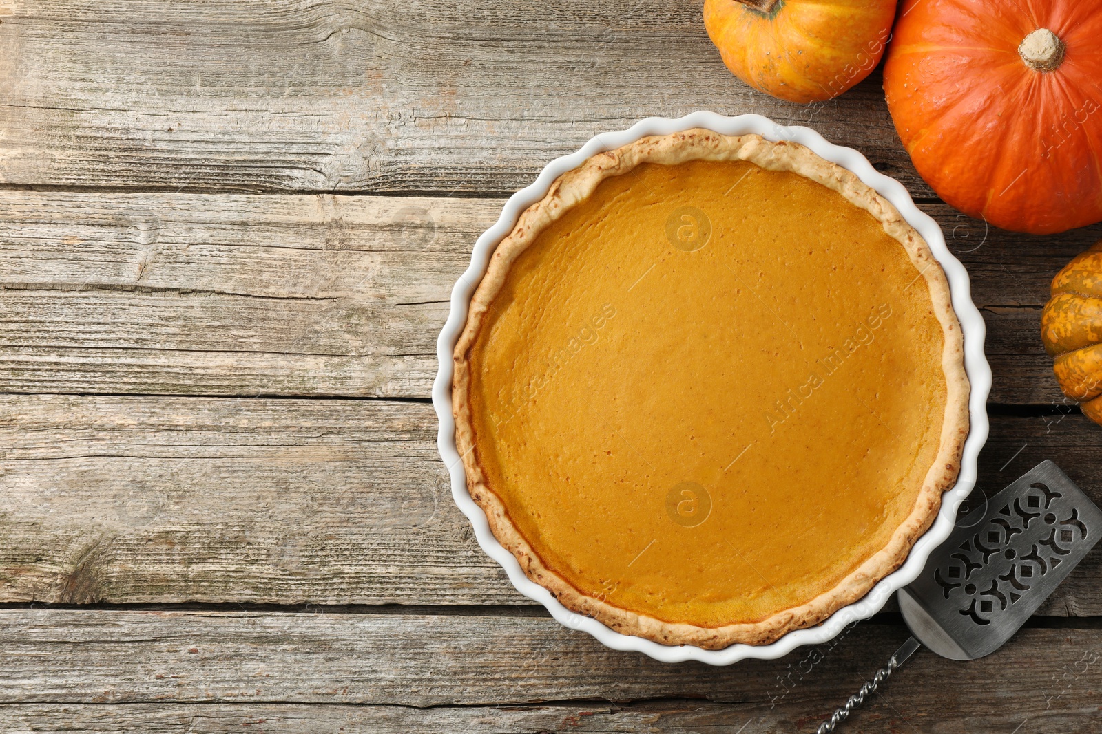 Photo of Tasty homemade pumpkin pie in baking dish, fresh pumpkins and cake server on wooden table, flat lay. Space for text