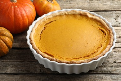 Tasty homemade pumpkin pie in baking dish and fresh pumpkins on wooden table, closeup