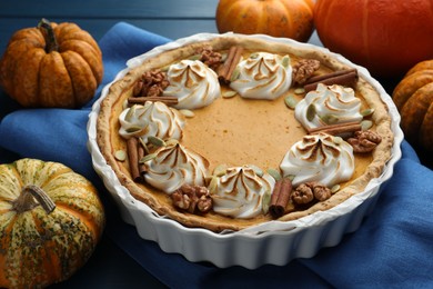 Photo of Delicious homemade pumpkin pie in baking dish and fresh pumpkins on blue wooden table, closeup