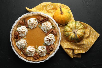 Photo of Delicious homemade pumpkin pie in baking dish and fresh pumpkins on black textured table, flat lay